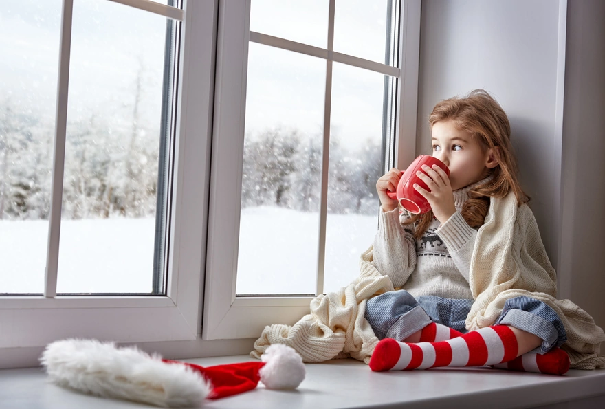 The girl wrapped blanket sits on the windowsill, drinking from a mug and looking out the window