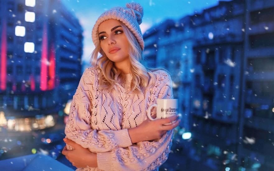 Блондинка с кружкой в руке, в тёплой кофте и шапке ёжится от холода