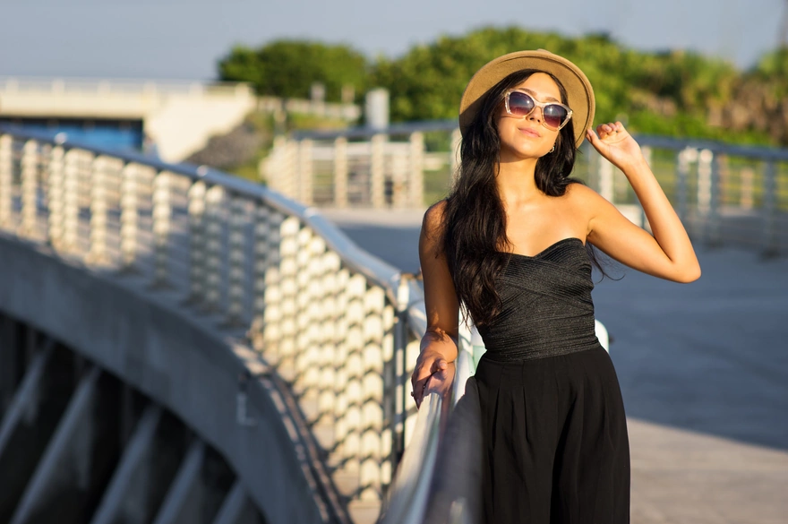 Стильная брюнетка в чёрном платье в шляпе и очках стоя на мосту позирует на закате солнца