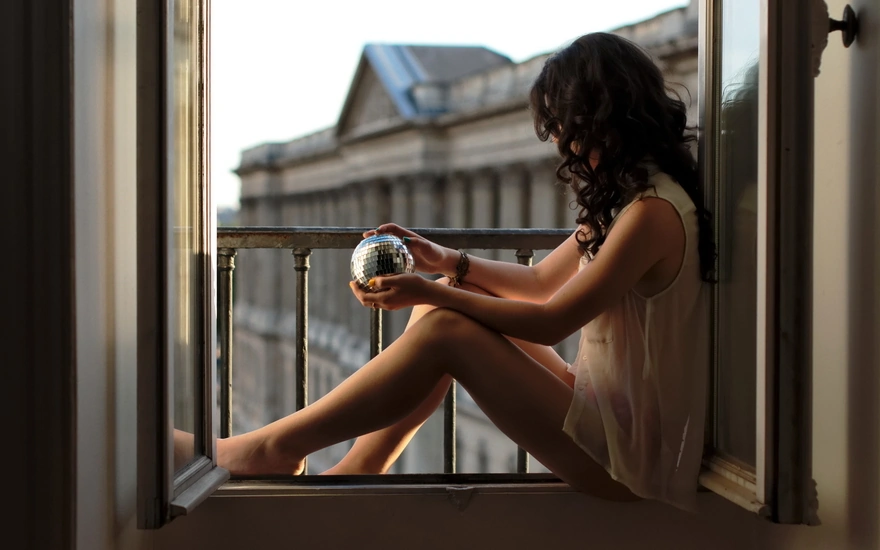 Брюнетка в блузке сидит на окне со стеклянным шаром в руках