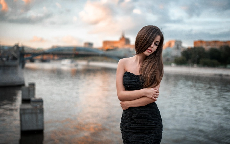 A girl in a black dress stands near the river, and behind her, a bridge is vaguely visible