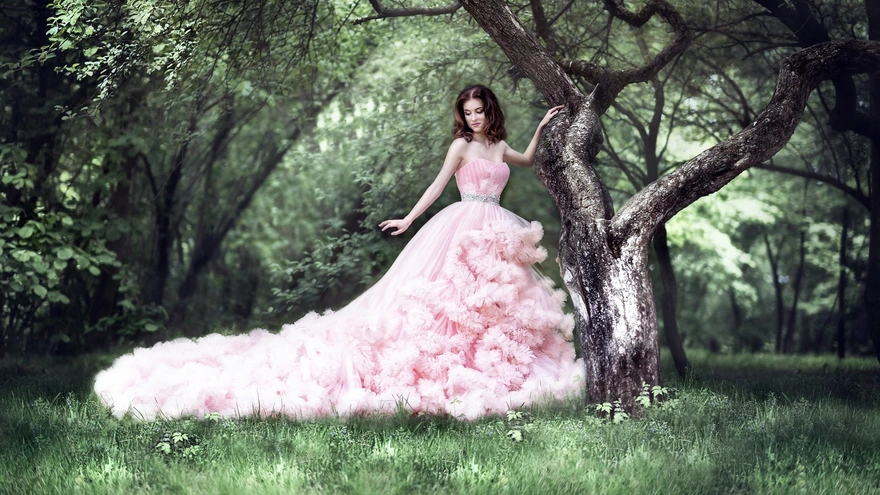 A girl in a lush pink dress with a train stands by a tree
