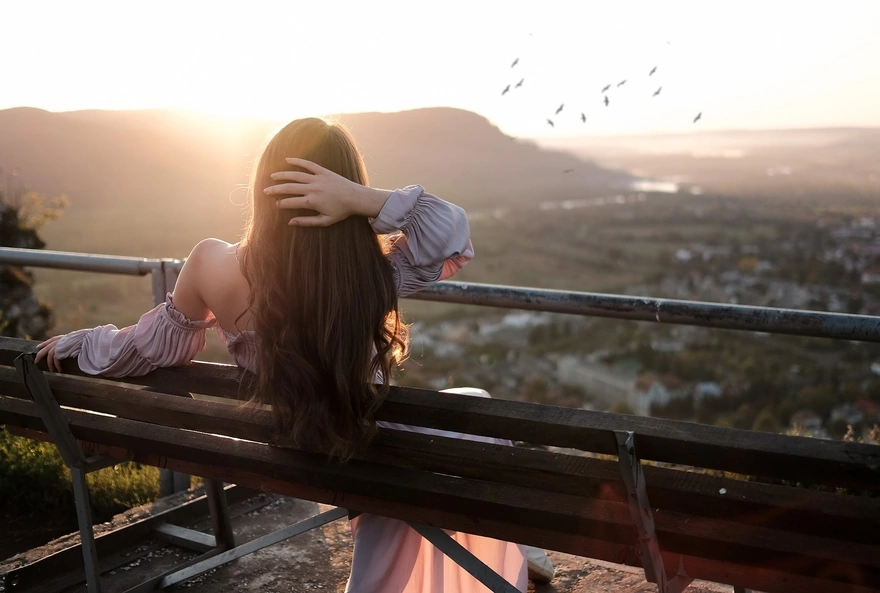 Девушка, сидя на скамейке и держа руку за головой, любуется закатом солнца