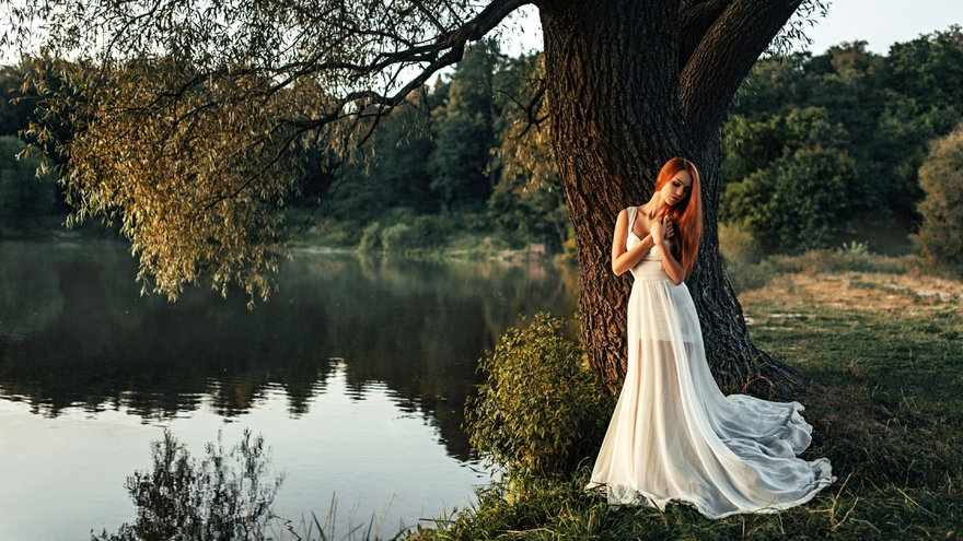 Длинноволосая рыжая девушка в белом длинном платье стоит у дерева