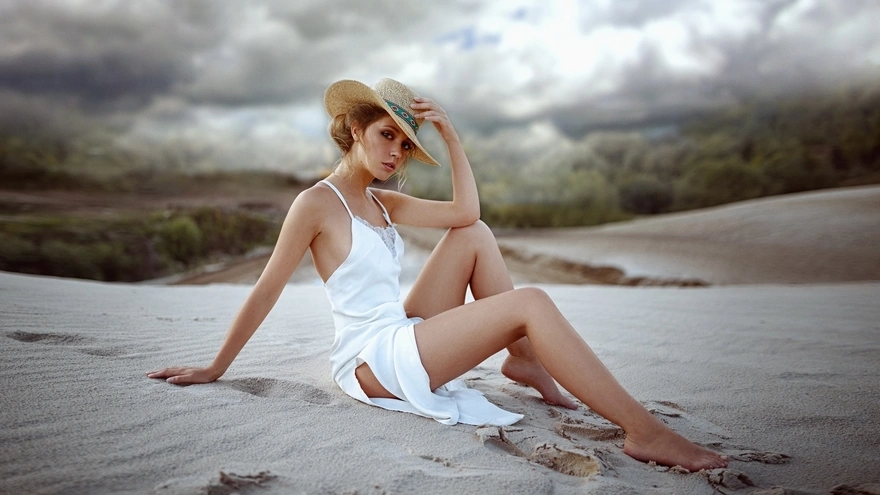 Girl sitting on the sand in a white sundress and a hat
