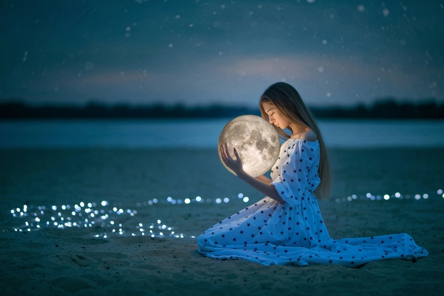 Девушка в платье сидит на песке и держит в руках сферу похожую на луну