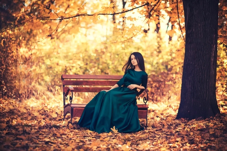 Брюнетка в длинном платье сидит на скамейке в осеннем парке