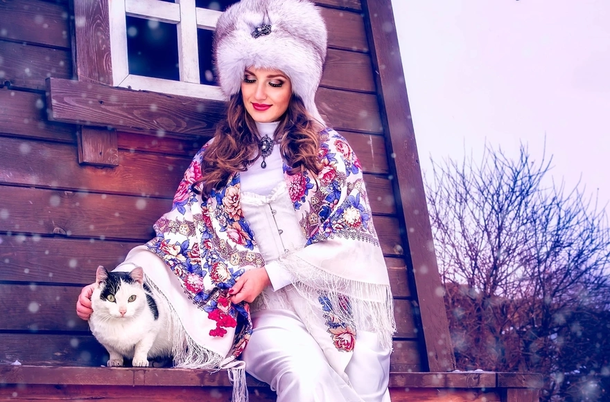 Девушка в меховой шапке и платке гладит кошку сидя у домика