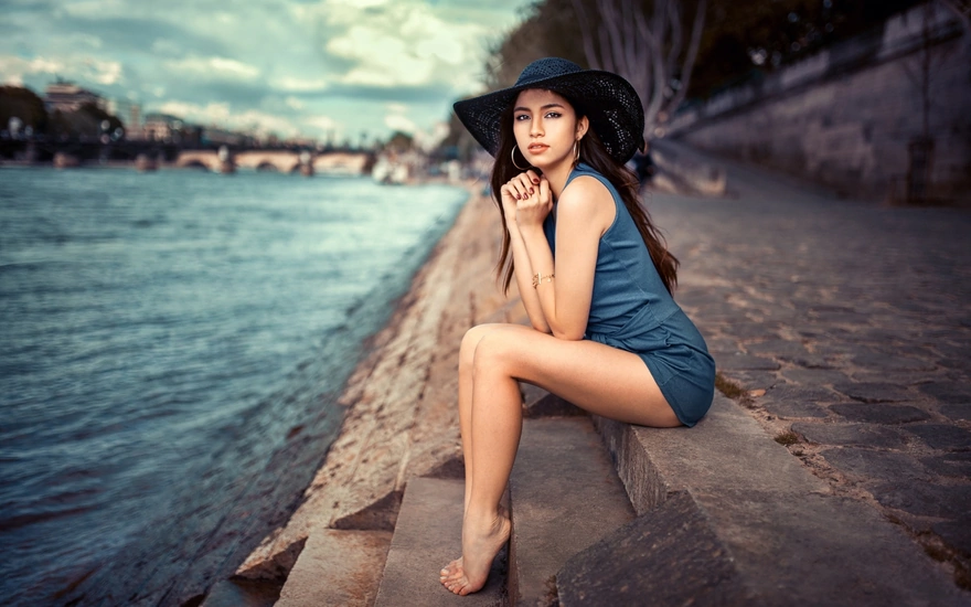 Брюнетка в синем платье и шляпе сидит на ступеньках на набережной у реки