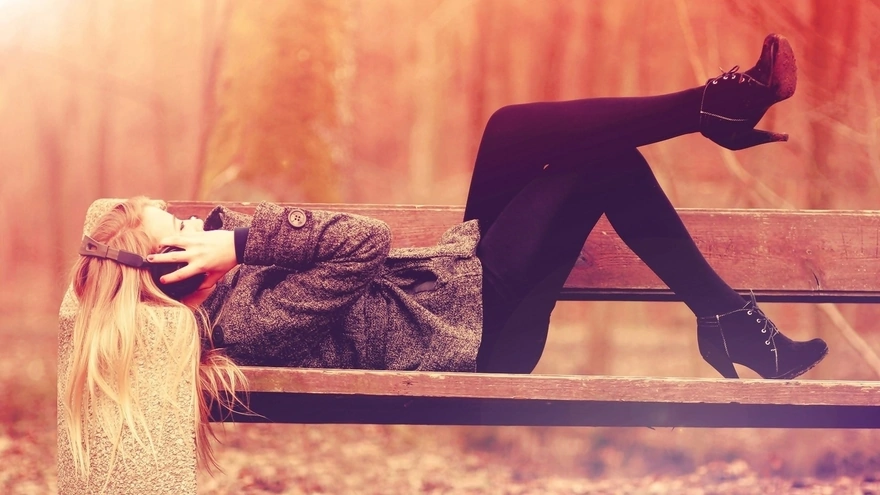 Девушка слушает музыку в наушниках лёжа на скамейке в парке