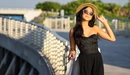 Картинка: Стильная брюнетка в чёрном платье в шляпе и очках стоя на мосту позирует на закате солнца