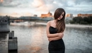 Image: A girl in a black dress stands near the river, and behind her, a bridge is vaguely visible