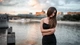 Картинка: Девушка в чёрном платье стоит возле реки, а позади неё расплывчато видно мост